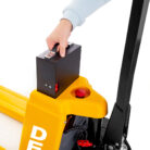Worker operating a yellow pallet jack, pressing a black control box mounted on the handle with a red emergency button nearby